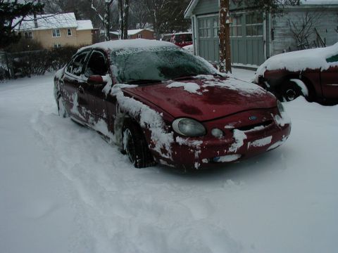 1997 Ford Taurus - A red Ford Taurus is stuck in deep snow, with snow covering its windshield and body. Another vehicle is visible in the background.