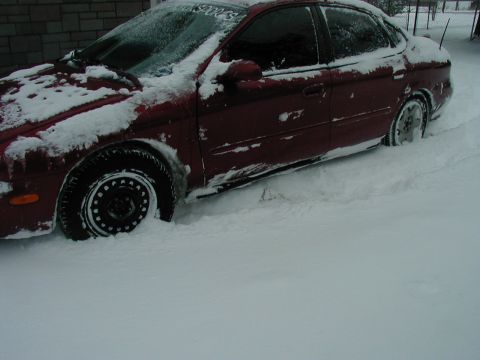 1997 Ford Taurus - A red sedan is partially buried in snow, with visible 'NASCAR' text on the windshield.