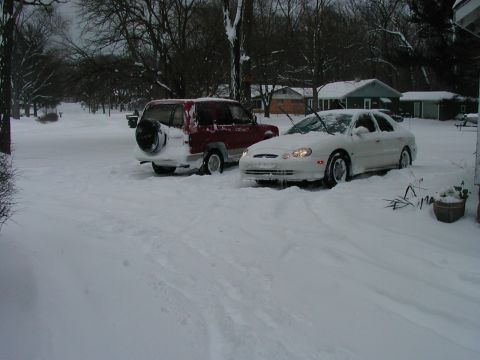 1997 Ford Taurus - Two vehicles are stuck in deep snow on a street in front of houses.
