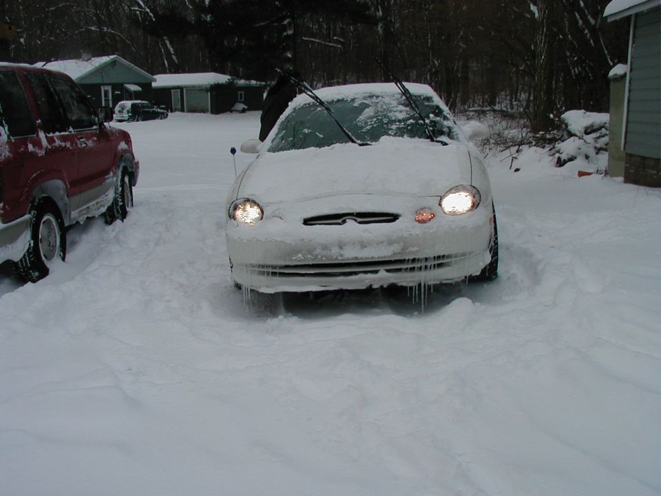 Front view of a white sedan in snow, icicles hanging from the bumper. A red SUV is parked to the left
