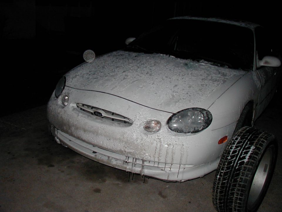 Front view of a white 1997 Ford Taurus covered in ice, with a spare tire next to it