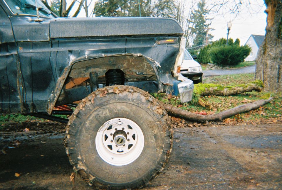 Lifted dark Ford truck on muddy off-road tires, front fender removed, in a yard
