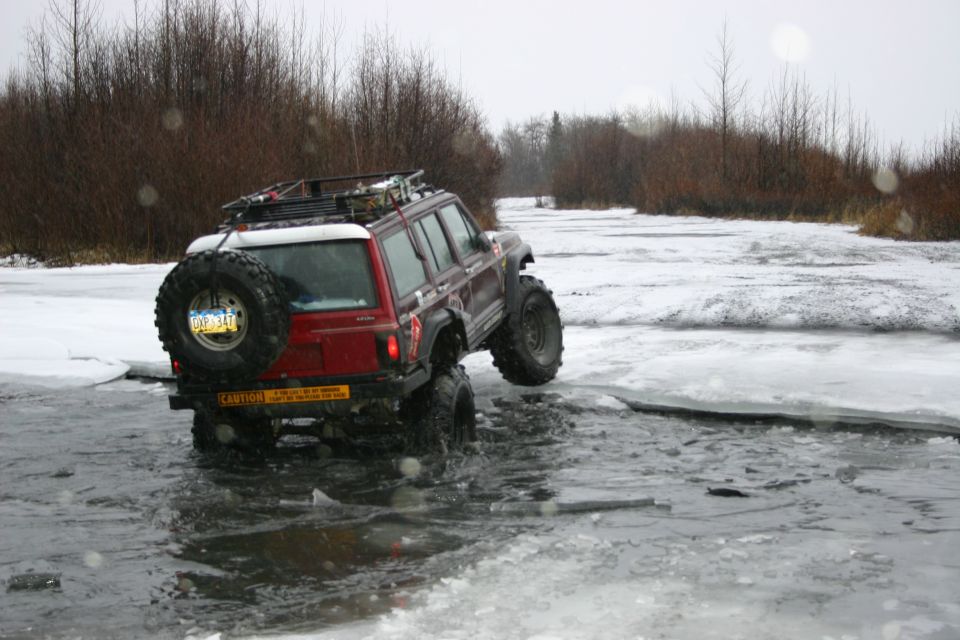 Red Jeep Cherokee XJ with oversized tires crossing a partially frozen body of water, spare tire mounted on rear door