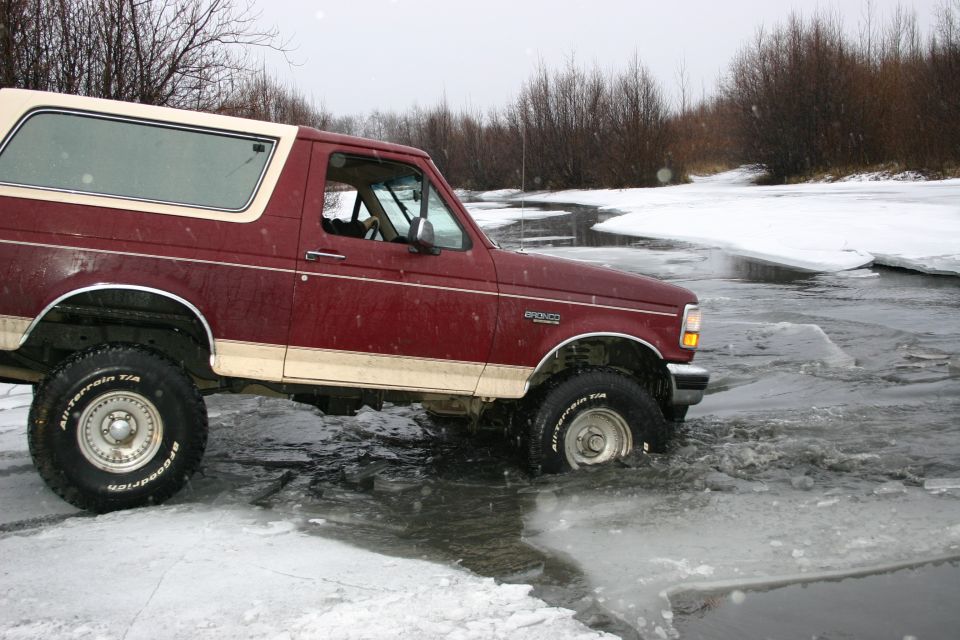 Maroon Ford Bronco with tan top driving through shallow icy water, trees and snow in background