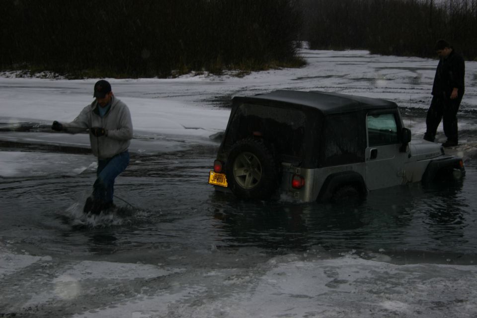 Man in water pulling a rope attached to a Jeep Wrangler stuck in icy water. Another person stands on the vehicle