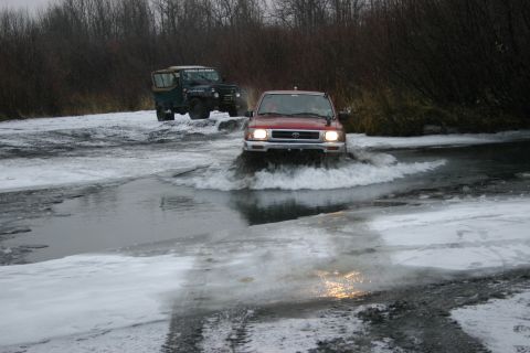 2003 Jeep TJ - Two off-road vehicles navigate a partially frozen river in a wooded area.