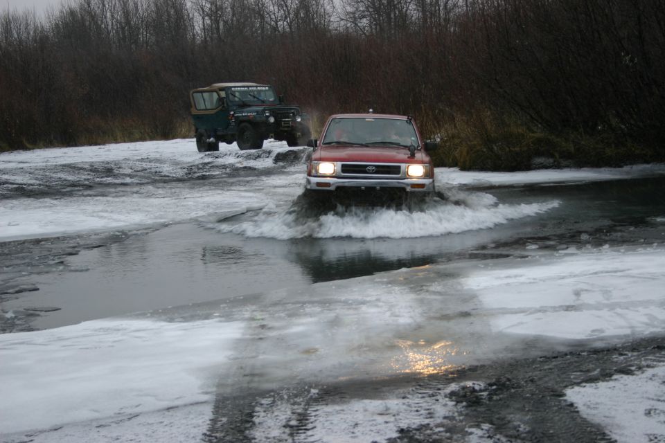 Red Toyota pickup truck driving through icy water, front view. Green Jeep behind