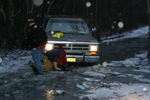 2003 Jeep TJ - A brown Dodge van is stuck in an icy, snowy road with two people working on it.