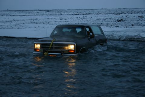 2003 Jeep TJ - A dark SUV is seen driving through a body of water, possibly a river or flooded area, with its headlights on.