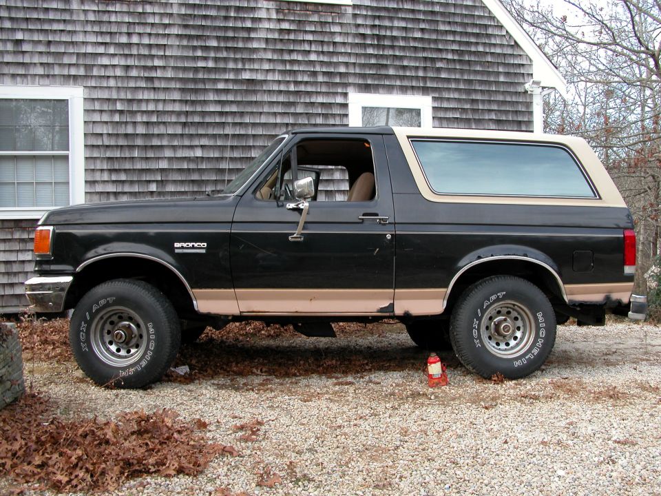 Black and tan Ford Bronco side view on gravel driveway by gray-shingled house, red bottle jack on ground