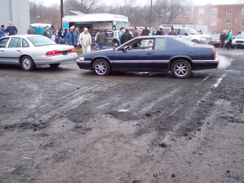 1991 Ford LTD Crown Victoria - A dark blue coupe is seen from the side, emitting smoke, with a silver sedan nearby in a lot.