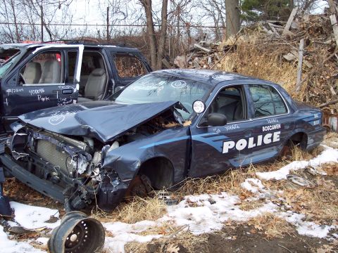 1991 Ford LTD Crown Victoria - A heavily damaged police car sits in a junkyard next to another vehicle, showing significant front-end damage.
