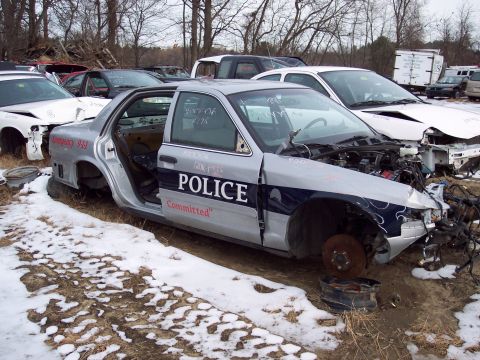 1991 Ford LTD Crown Victoria - A damaged police car sits in a snowy junkyard, stripped of parts and abandoned.