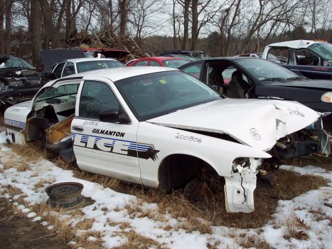 1991 Ford LTD Crown Victoria - A damaged white police car sits in a junkyard, surrounded by other vehicles and bare trees.