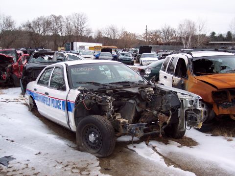 1991 Ford LTD Crown Victoria - A damaged Springfield Police car sits in a junkyard with other vehicles under a cloudy sky.