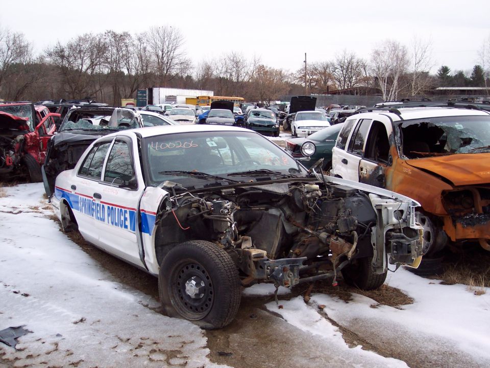Police car with 'Springfield Police' on door, missing hood and engine parts. Other wrecked cars in background