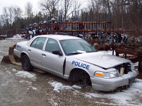 1991 Ford LTD Crown Victoria - A silver police car, marked with 'POLICE' and '911', sits in a junkyard with its wheels removed.