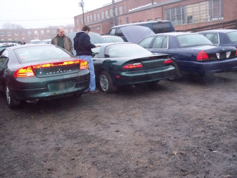 1991 Ford LTD Crown Victoria - Two cars, a dark green coupe and a blue sedan, are parked side-by-side in a lot with muddy ground.