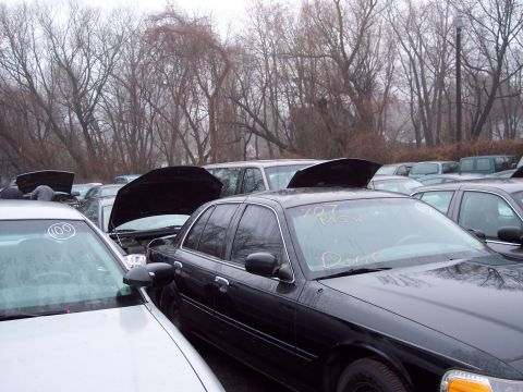 1991 Ford LTD Crown Victoria - A black sedan sits in a junkyard with its hood open, surrounded by other cars under a cloudy sky.