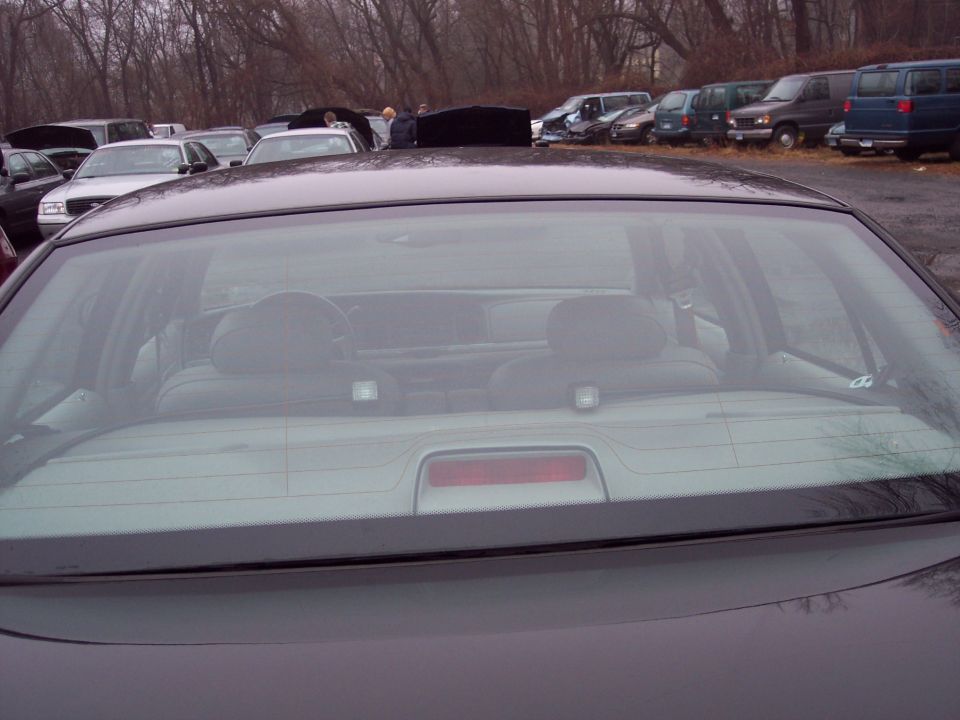 Rear interior view through a sedan's back window, showing headrests and steering wheel. Multiple vehicles parked in background