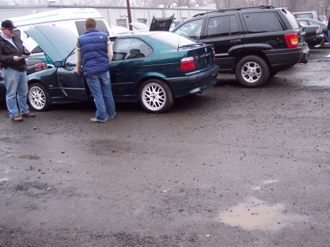 1991 Ford LTD Crown Victoria - Two men inspect a green BMW coupe parked next to a black SUV in a muddy lot.