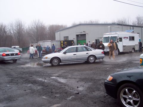 1991 Ford LTD Crown Victoria - A silver sedan is parked on a muddy lot next to a building, with several people standing around.