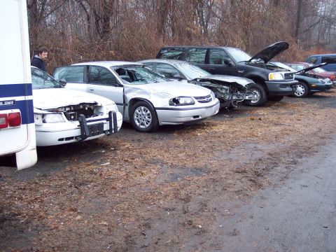 1991 Ford LTD Crown Victoria - A line of damaged vehicles, including a white 1991 Ford Crown Victoria LTD, are parked on a wet, debris-strewn lot.