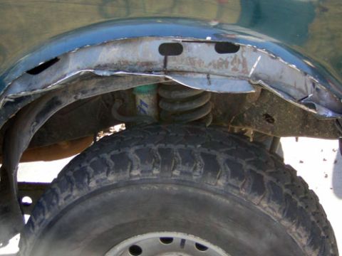 1995 Ford Bronco - Close-up of a dark green truck's front wheel well with the fender lip freshly cut back for tire clearance, leaving raw trimmed sheet-metal…
