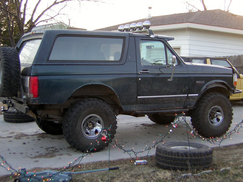 Lifted dark green full-size Bronco in a driveway strung with Christmas lights at dusk