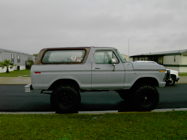 Lifted 1978 Ford Bronco parked on a street, side view. Another white vehicle is visible behind it