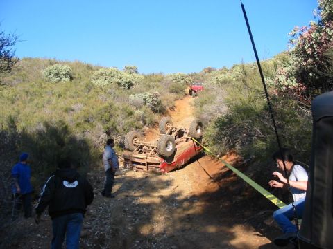 1990 Ford Bronco - red vehicle rolled onto its roof on a rutted dirt off-road trail in green hills, with bystanders and a tow strap nearby.