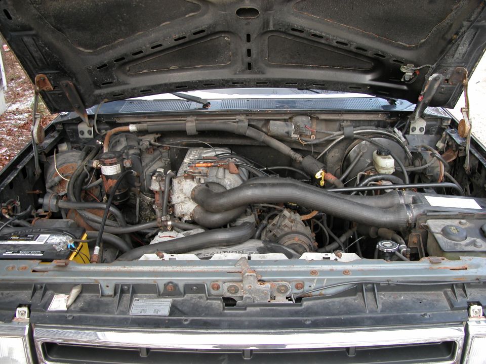 Open engine bay of a truck showing inline engine, intake hose and rusty radiator support