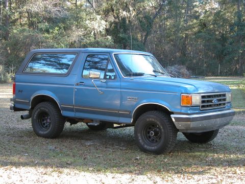 1990 Ford Bronco - A blue Ford Bronco is parked on a grassy area with trees in the background.