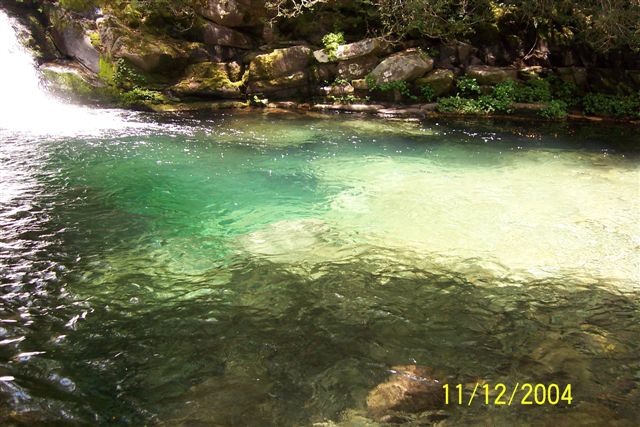 waterfall cascading into a clear, shallow river with rocks and trees along the bank. Supermotors.net logo visible
