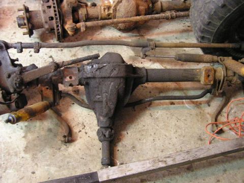 1988 Ford Bronco - Rusty vehicle axle and suspension parts are laid out on a concrete floor, possibly for repair or modification.