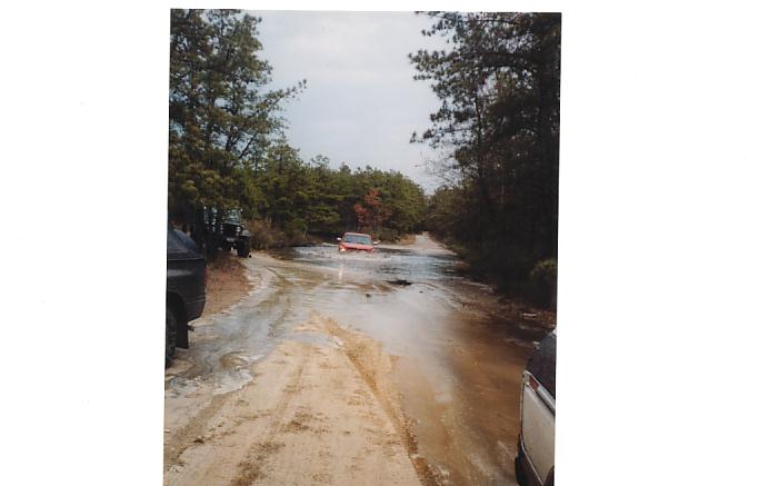 Red SUV driving through a muddy, flooded dirt road with trees on either side. Another vehicle is partially visible on the right