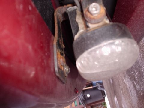 1998 Ford Taurus - Tight low-angle shot along a dark red body panel at a rubber bump stop or hood/trunk stop with a corroded steel bracket and rusty bolt head…
