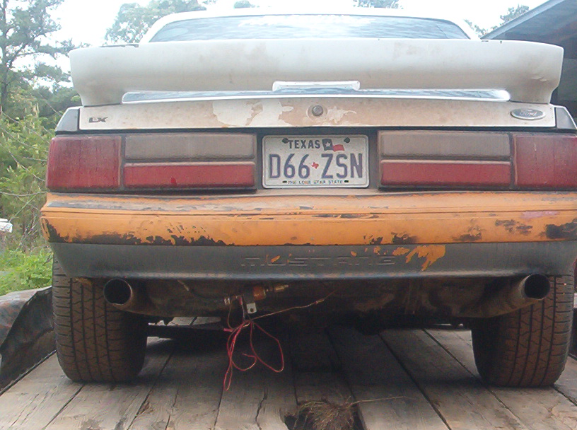 Rear of white 1989 Ford Mustang LX with orange bumper and Texas plate D66-ZSN on a wooden trailer