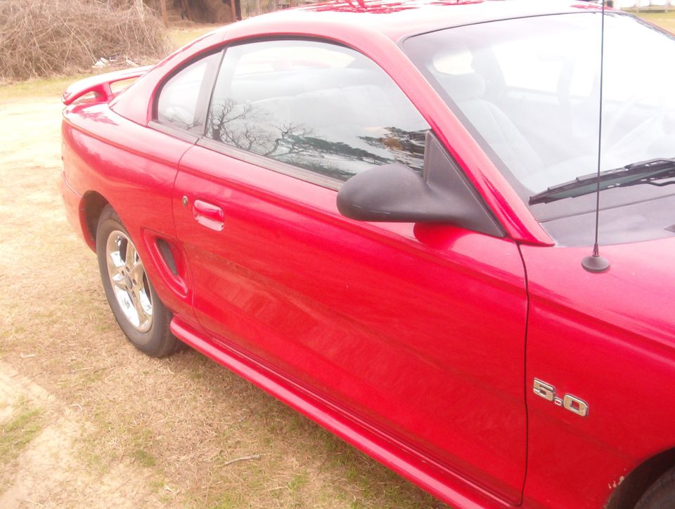 Red Ford Mustang coupe passenger side with chrome wheel and 5.0 badge, parked on dirt.