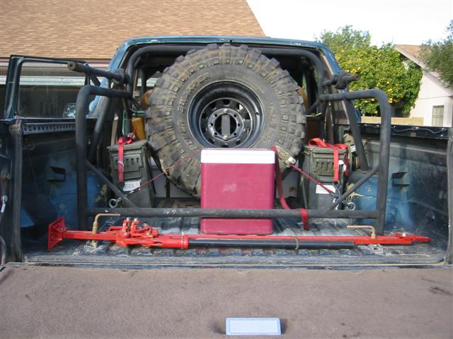 Truck bed with spare tire on roll cage, ammo cans, red high-lift jack and cooler
