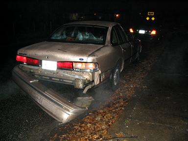 1999 Ford Crown Victoria - A damaged 1990s Ford Crown Victoria is seen from the rear, its bumper hanging off, on a dark, wet road.
