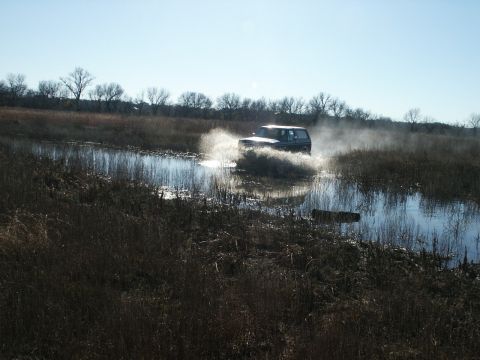 1991 Ford Bronco - Backlit wide shot of a full-size SUV with a light-colored hardtop running through standing water in a flooded winter marsh, kicking up a br…