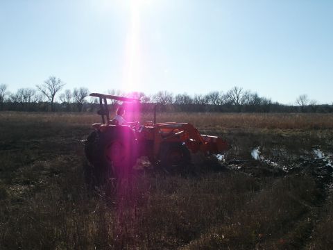 1991 Ford Bronco - A person operates an orange tractor with a front loader in a muddy, grassy field under a bright sun.