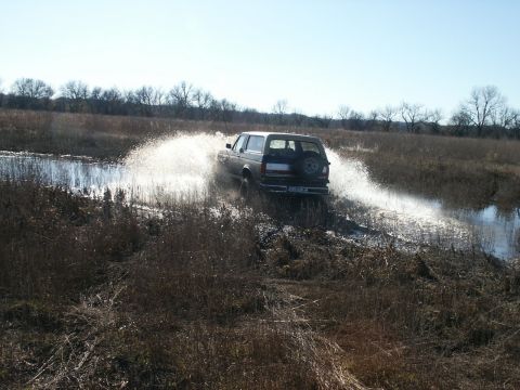 1991 Ford Bronco - Rear three-quarter view of a silver/tan full-size Ford Bronco charging through a shallow marsh pond, kicking a tall curtain of water off bo…