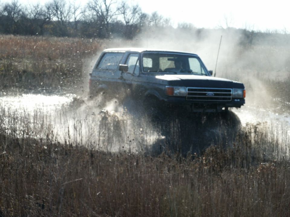 Dark blue Ford Bronco driving through muddy water, kicking up spray. The vehicle is viewed from the front-right