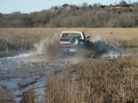 1991 Ford Bronco - Rear view of a Ford Bronco with a white hardtop and a tailgate-mounted spare on a white spoke wheel plowing through a flooded field, throwi…