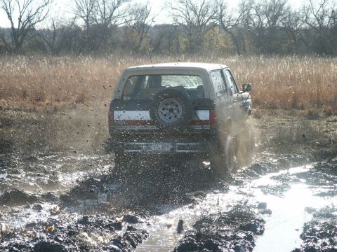 1991 Ford Bronco - Rear view of a dark full-size Ford Bronco with a white hardtop running through a churned mud bog, slinging clods and spray from the rear ti…