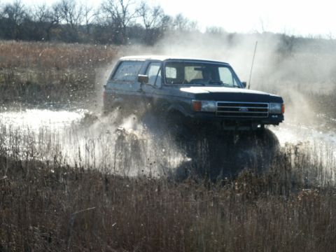 1991 Ford Bronco - Three-quarter front action shot of a dark Ford Bronco with a white hardtop charging through a flooded, weed-choked field and kicking up she…