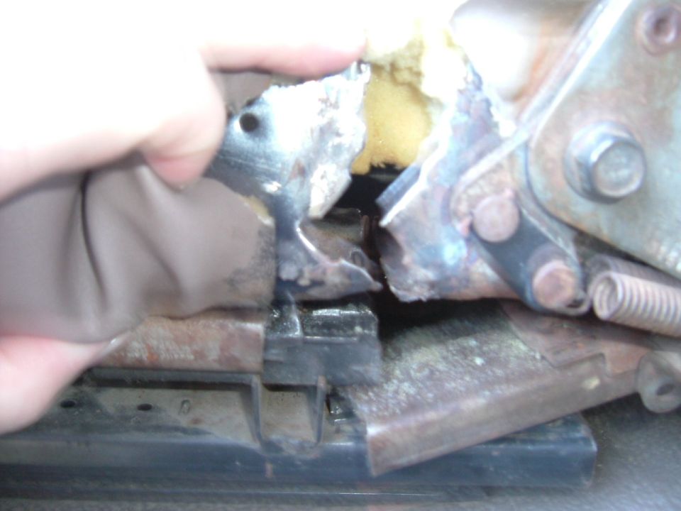 Hand lifting seat upholstery to reveal a rusted, cracked seat frame and track with exposed foam