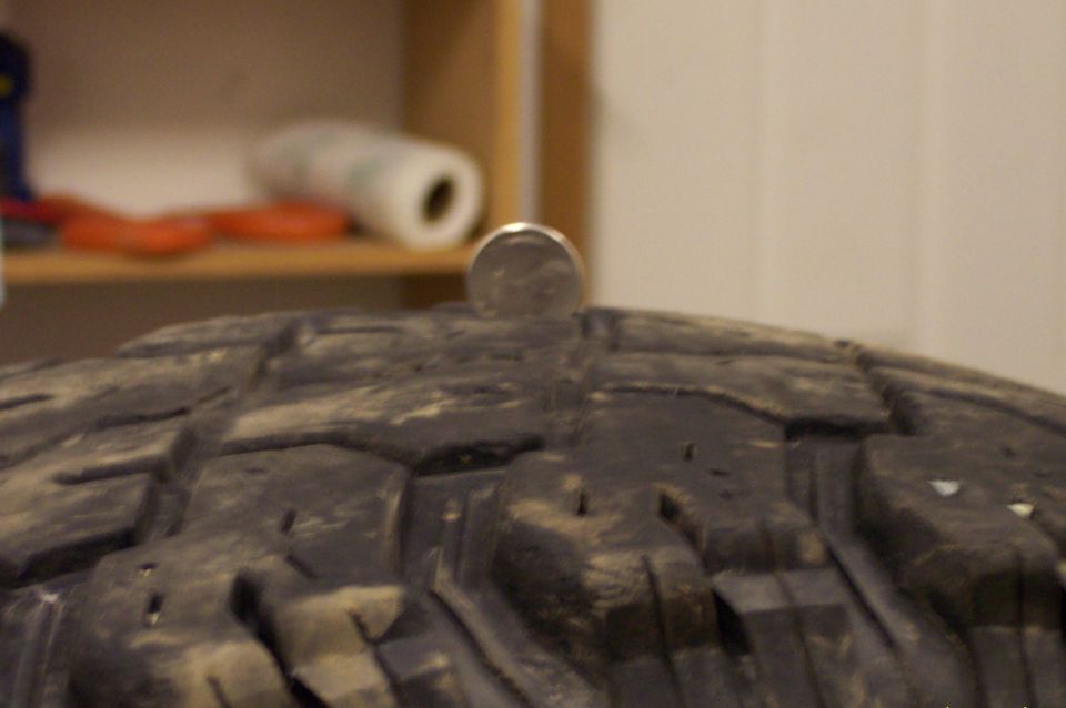 Close-up of a tire tread with a quarter balanced on its surface. Background shows a shelf with items